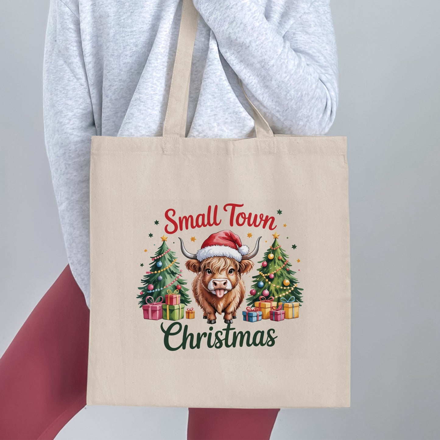 Tote bag with highland cow wearing Santa hat, Christmas trees, and gifts. Text: Small Town Christmas.