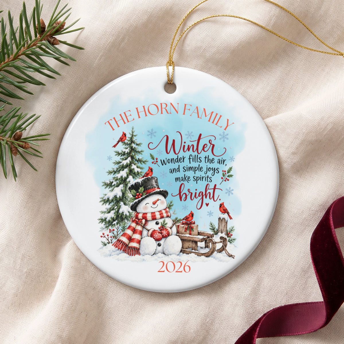 Round white ceramic Christmas ornament featuring a snowman, winter trees, red cardinals, and text reading 2026.