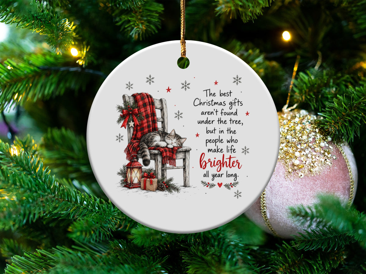 Round white Christmas ornament featuring a sleeping cat on a plaid blanket with holiday text hanging on a tree.
