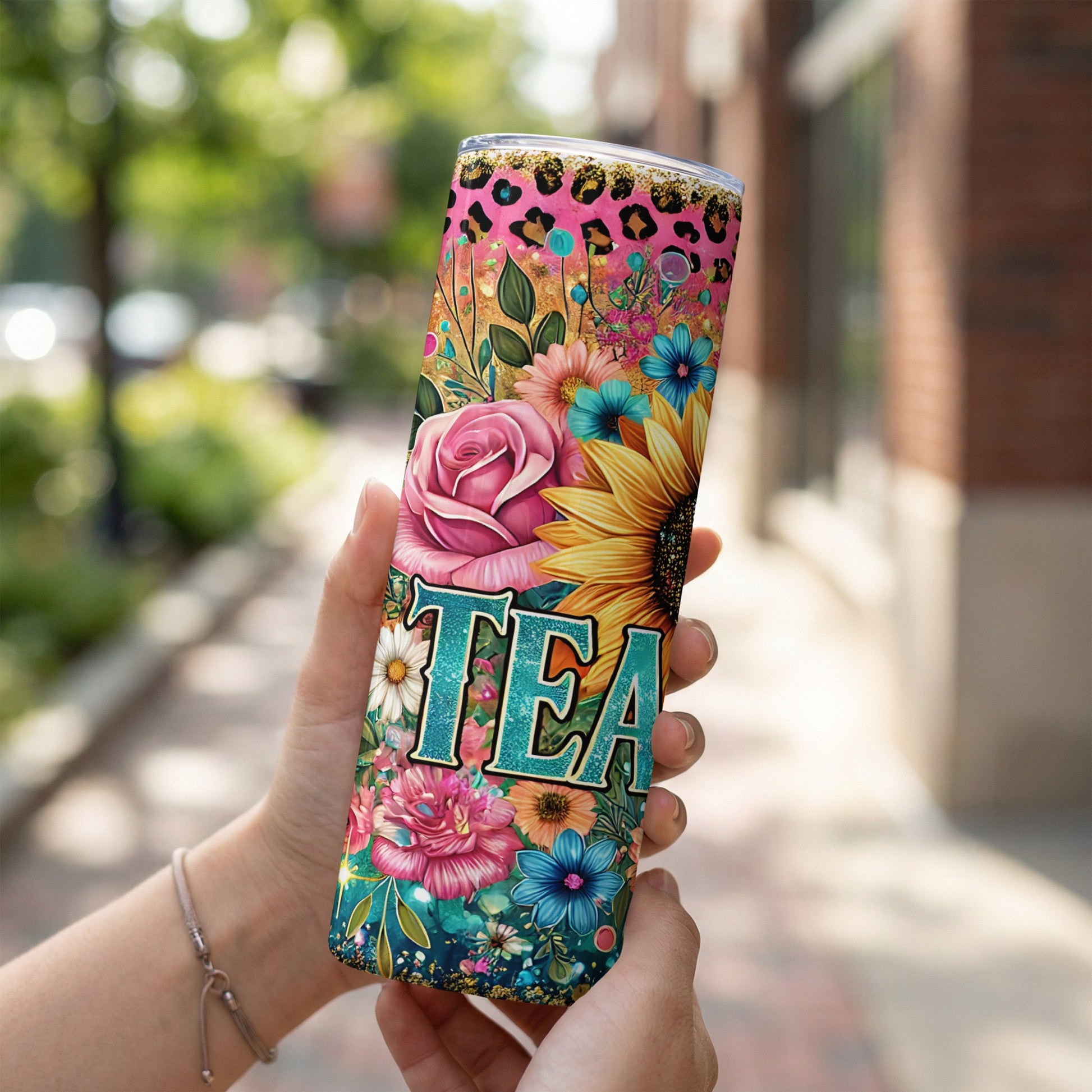 Tumbler with floral pattern and "TEA" text held by hands outdoors.