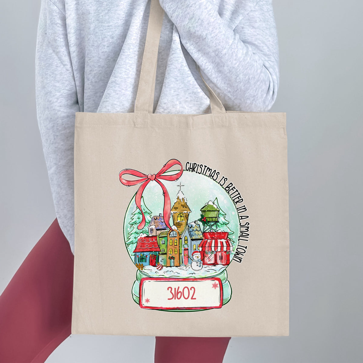 Tote bag featuring a Christmas snow globe with a small town scene, text: "Christmas is better in a small town" and "31602".