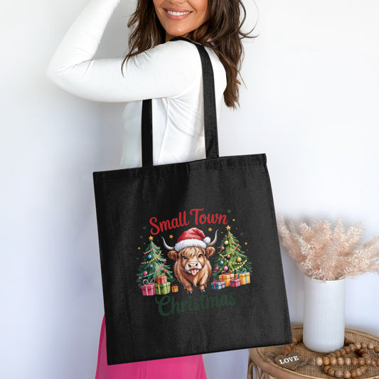Black tote bag with "Small Town Christmas" text and a Highland cow wearing a Santa hat.