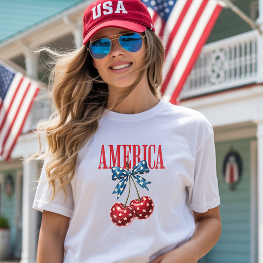 Woman wearing a white t-shirt with 'AMERICA' and cherry design, red cap, sunglasses, in front of American flags.
