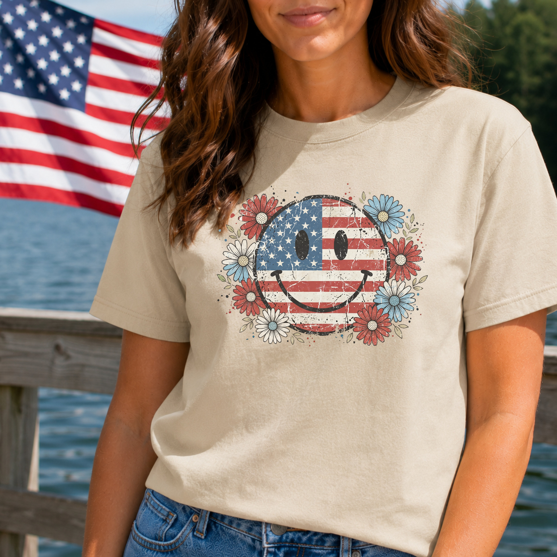 Person wearing a t-shirt with a smiley face design featuring an American flag and flowers, standing in front of an American flag and water.