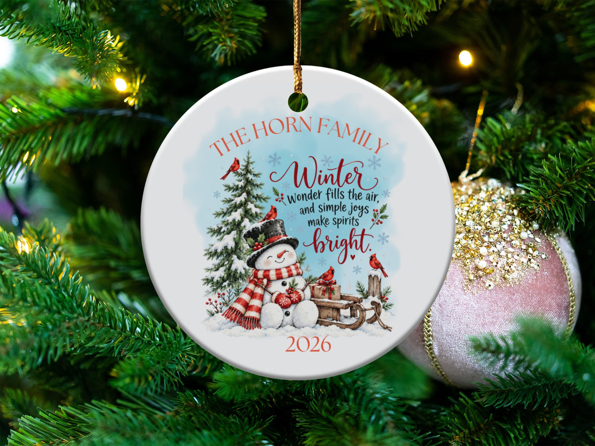 Round white ceramic ornament hanging on a Christmas tree, featuring a snowman, pine trees, red cardinals, and the text The Horn Family 2026.