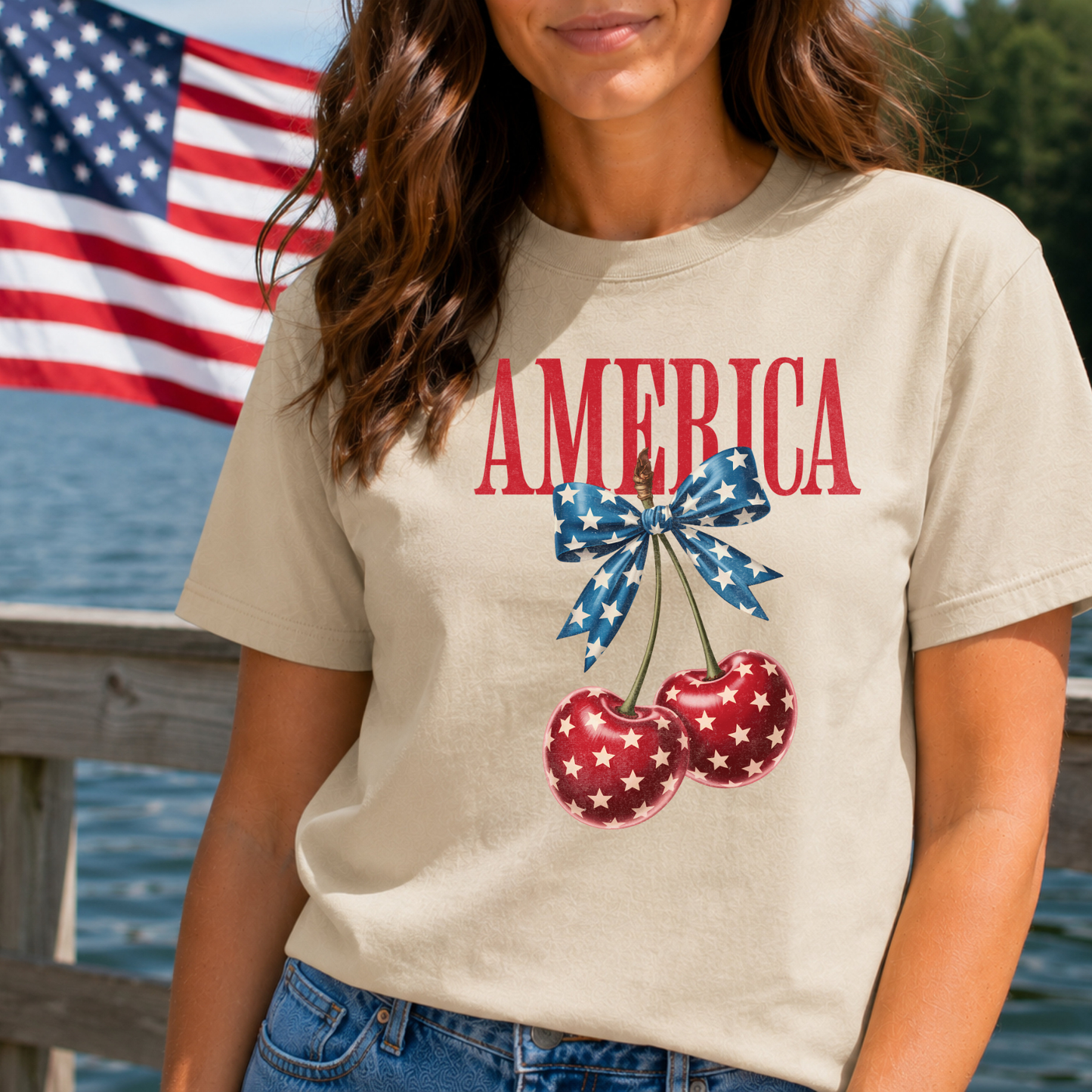 Person wearing a t-shirt with 'AMERICA' and cherry design, standing by a body of water with an American flag in the background.