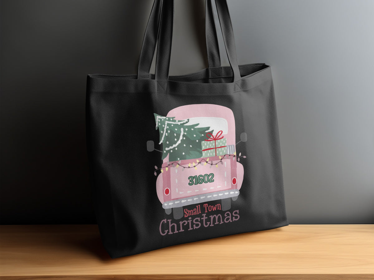 Black tote bag with "Small Town Christmas" design featuring a pink truck carrying a Christmas tree and presents.