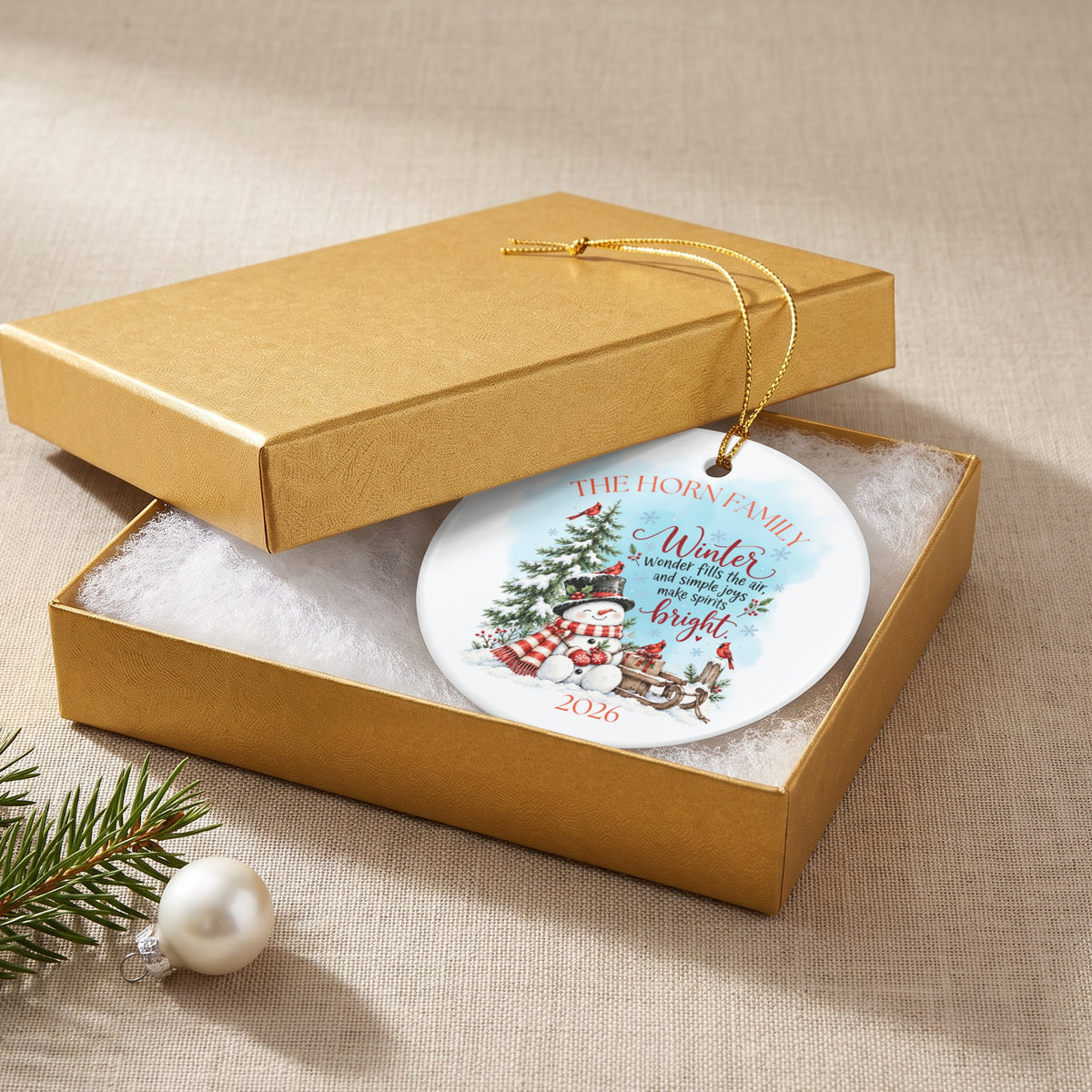 Personalized round white Christmas ornament in a gold gift box with a snowman, pine tree, and winter text.
