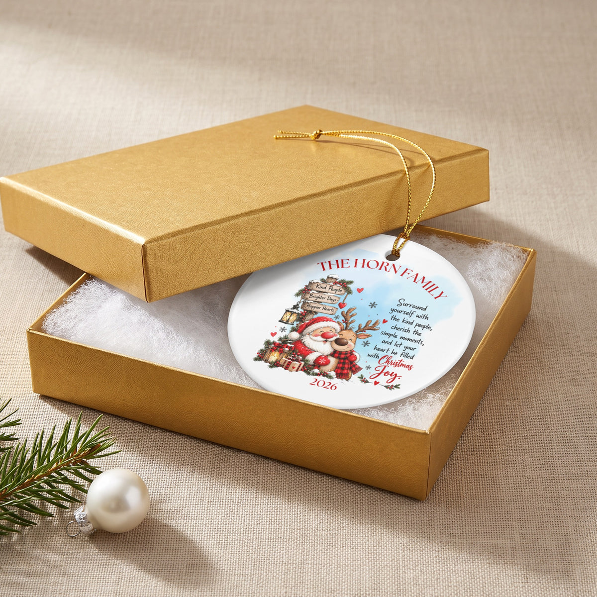 A round white Christmas ornament with holiday graphics and family text inside an open gold gift box.