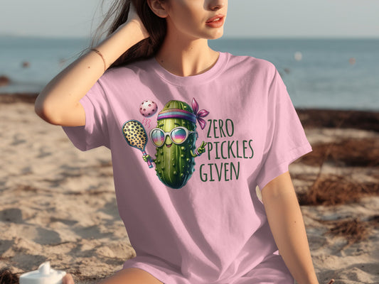 Pink t-shirt with a cartoon pickle wearing sunglasses, holding a tennis racket and ball.