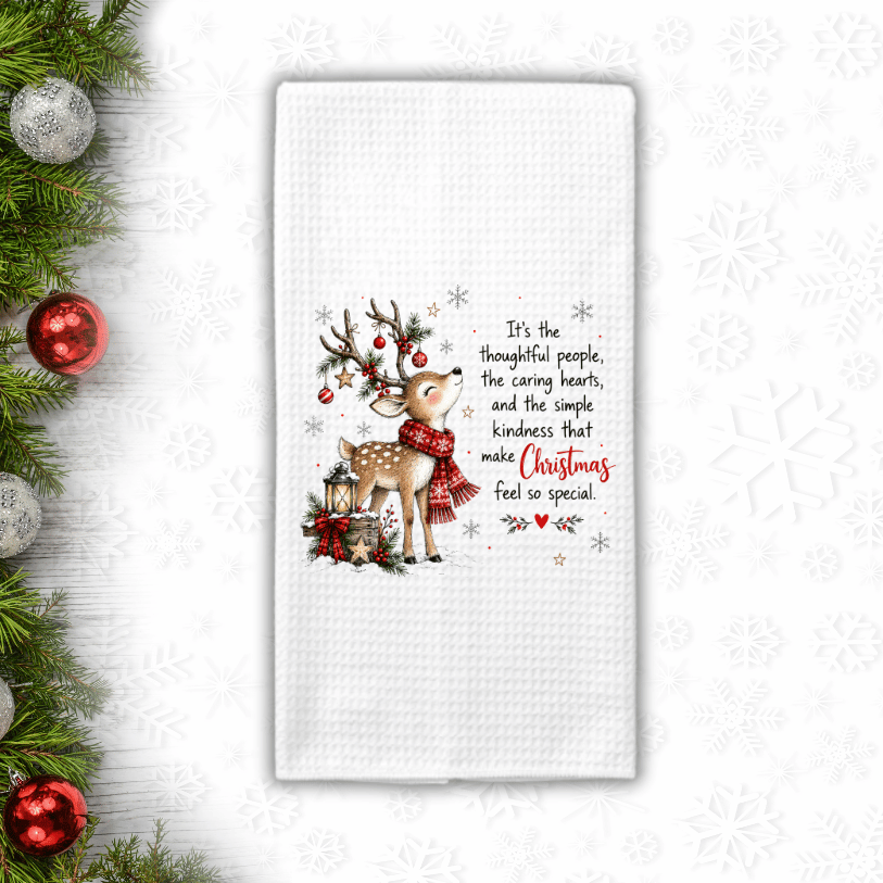 A white waffle-weave kitchen towel with a festive Christmas design featuring a deer, ornaments, and a quote, displayed on a white background with holiday greenery and ornaments in the corner.