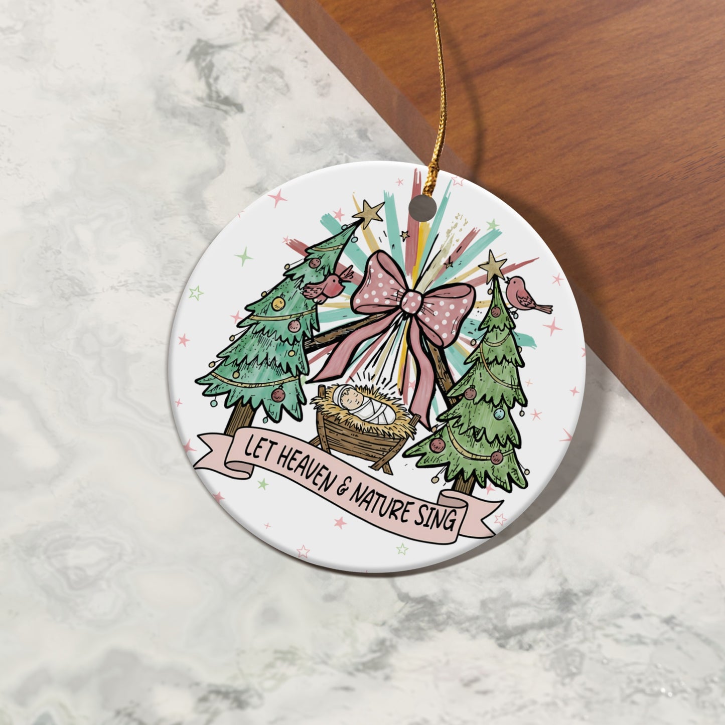 Christmas ornament with nativity scene, trees, pink bow, and text "LET HEAVEN & NATURE SING".