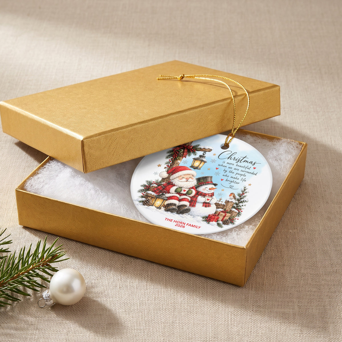 A round ceramic Christmas ornament featuring Santa and a snowman rests inside an open gold gift box.
