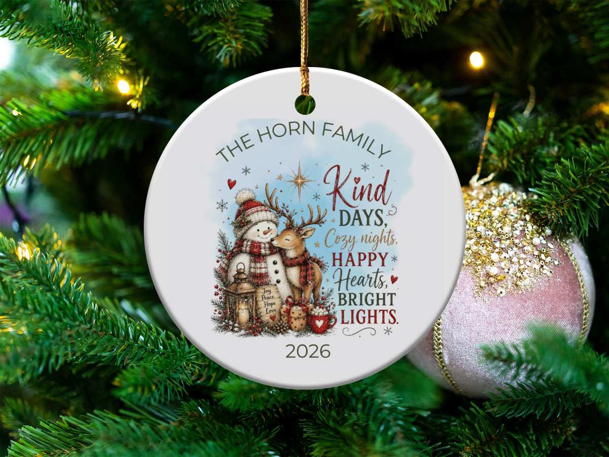 A round white ceramic Christmas ornament hanging on a tree features a snowman and reindeer illustration with personalized text.