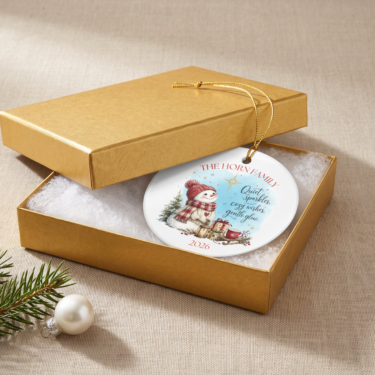 A round white ceramic Christmas ornament featuring a snowman graphic resting inside an open gold gift box.
