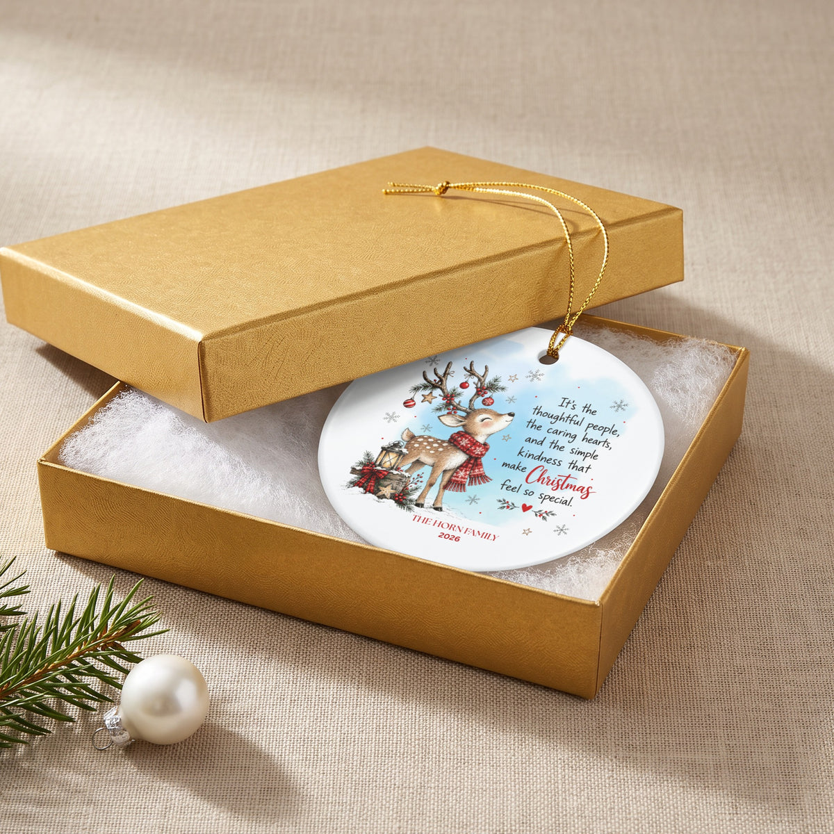 A round white Christmas ornament with a reindeer design inside a gold gift box.