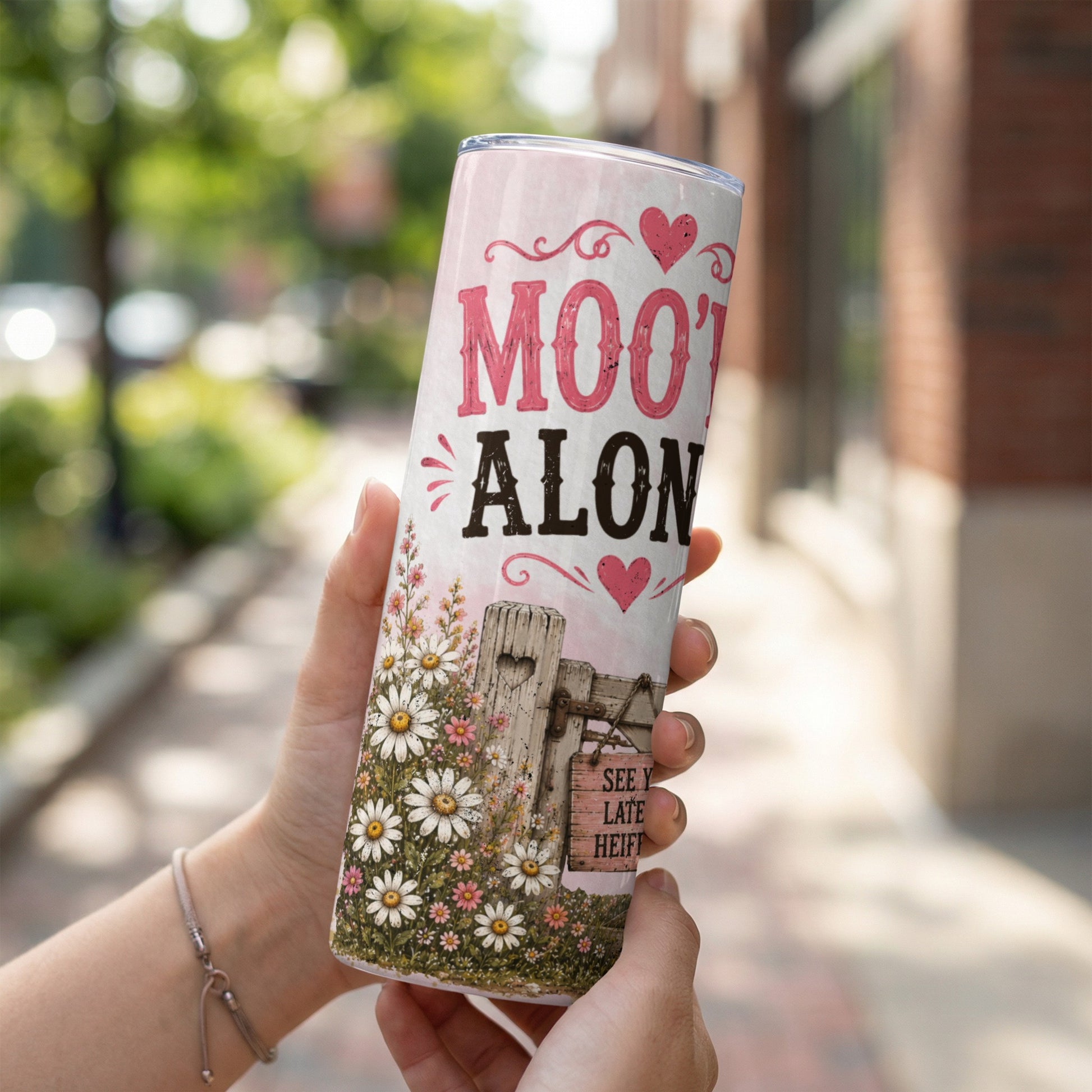 Tumbler with "Moo't Alone" text and daisies on a rustic fence background.