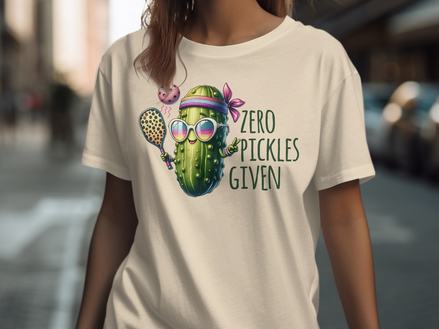 T-shirt with pickle character wearing sunglasses holding pickleball paddle and ball.