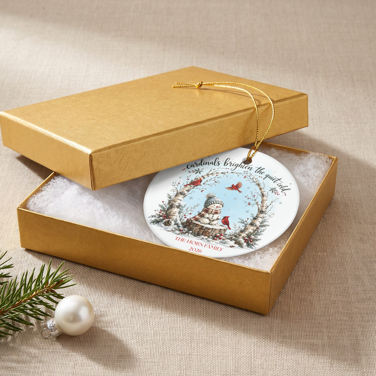A round ceramic Christmas ornament featuring a snowman and red cardinals lies inside an open gold gift box.